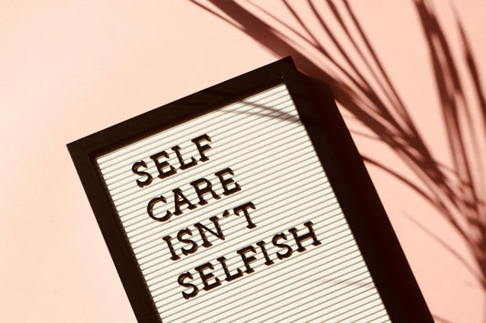 Ein Letterboard mit der Botschaft ‚Self Care Isn’t Selfish‘, fotografiert auf einem zartrosa Hintergrund mit Schatteneffekten. Ein inspirierendes Bild zur Betonung von Selbstfürsorge und Achtsamkeit