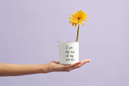 I am the hero of my own life- HaEine gelbe Blume, die aus einer weißen Tasse mit der Aufschrift 'I am the hero of my own life' wächst. Die Tasse wird von einer ausgestreckten Hand vor einem zarten lila Hintergrund gehalten.